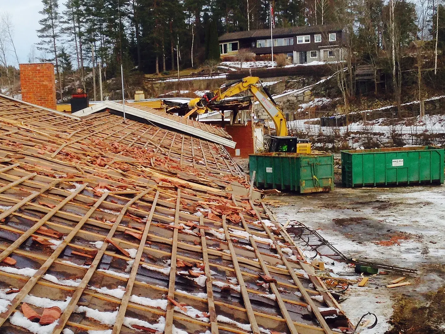 Riving av tak på en bygning, med en gul gravemaskin i arbeid og avfallskontainere i grønt i bakgrunnen.