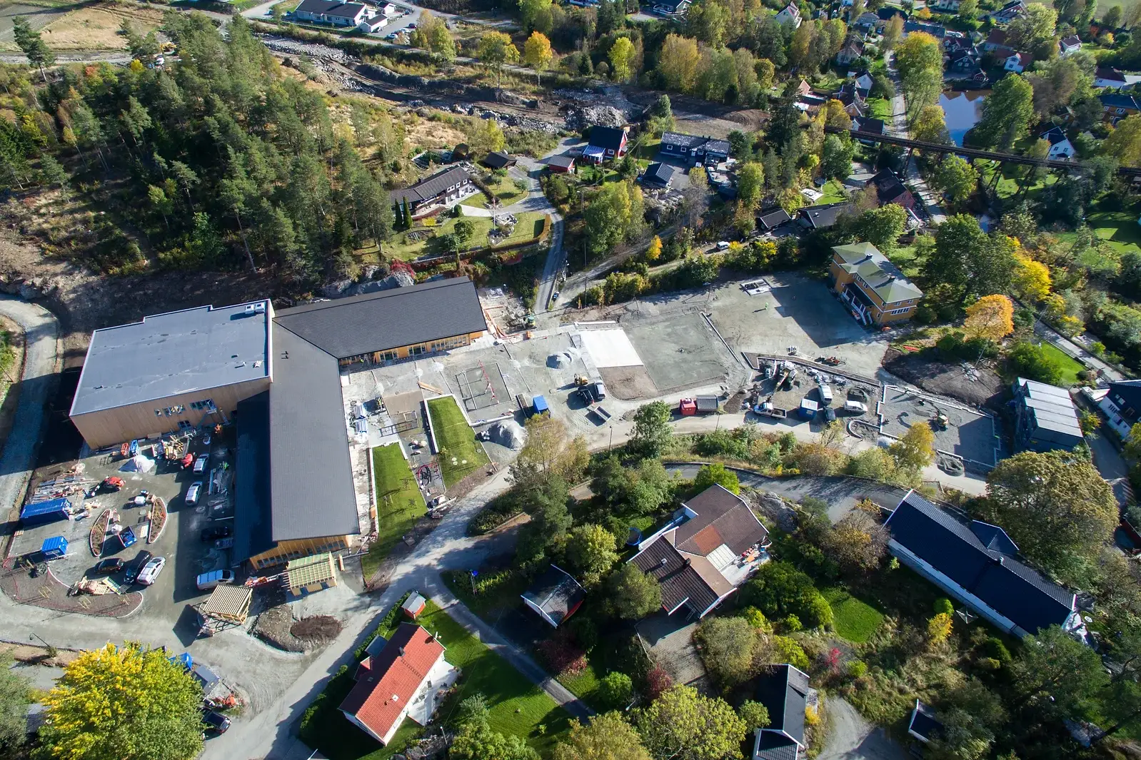 Fugleperspektiv av en skolegård under konstruksjon omgitt av grøntområder og hus, med flere ferdige bygningsstrukturer.